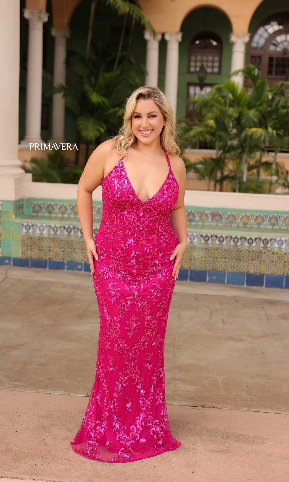 A woman stands outdoors, smiling in front of a tiled wall and greenery, wearing the Primavera Plus-Size Long Beaded Prom Dress 14043 in bright pink with thin straps.